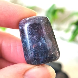 May include: A polished, dark blue-purple gemstone with a rounded rectangular shape. The stone has a smooth, reflective surface with subtle variations in colour and texture. The gemstone is held between two fingers.