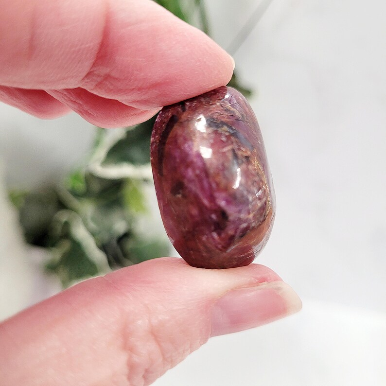 Puede incluir: Una gema pulida de forma ovalada, de un tono burdeos intenso, con vetas marrones y tonos rojos m&aacute;s claros. La piedra se sostiene entre dos dedos, mostrando su superficie lisa y reflectante. La gema parece ser un mineral natural.