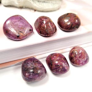 May include: Six polished, oval gemstones in deep red and purple hues. The stones have a glossy finish with varied colour patterns and textures. They are arranged on a white surface.