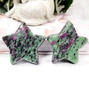 May include: Two star-shaped gemstones with a green base colour, speckled with black and magenta. The gemstones are displayed on a white surface, with a blurred background featuring a white statue and greenery.