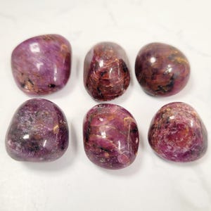 Puede incluir: Seis piedras preciosas pulidas y rodadas en tonos rojo intenso y p&uacute;rpura. Las piedras tienen una forma suave y redondeada y un acabado brillante, con variaciones naturales en color y textura. Las piedras est&aacute;n dispuestas sobre una superficie blanca.