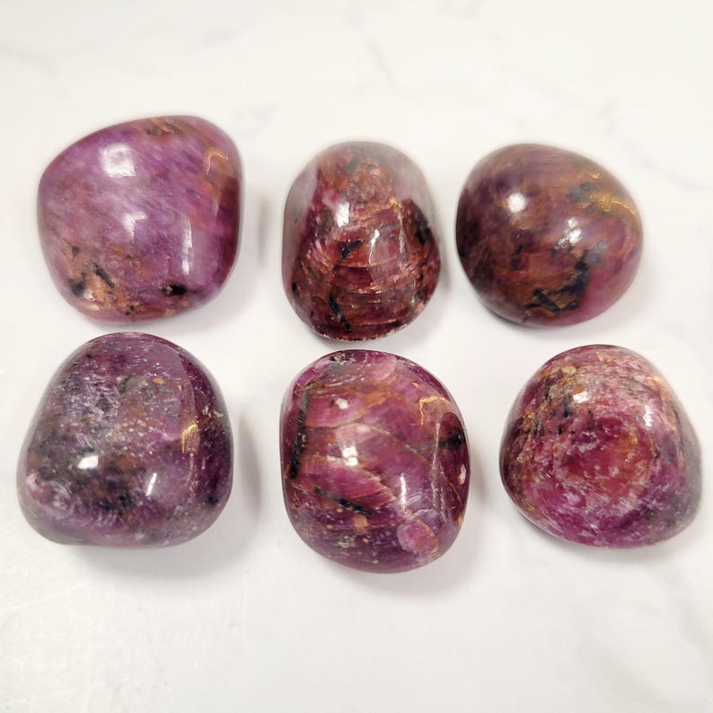 Puede incluir: Seis piedras preciosas pulidas y rodadas en tonos rojo intenso y p&uacute;rpura. Las piedras tienen una forma suave y redondeada y un acabado brillante, con variaciones naturales en color y textura. Las piedras est&aacute;n dispuestas sobre una superficie blanca.