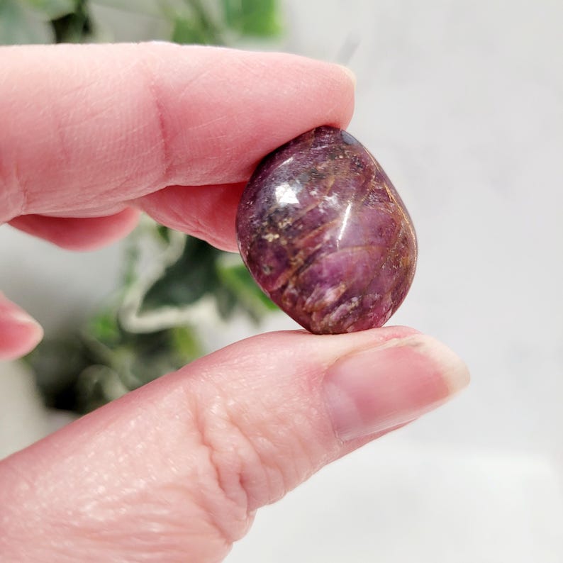 Puede incluir: Una gema pulida, de forma ovalada, con un tono burdeos intenso, acentuada por vetas marrones y toques blancos. La piedra se sostiene entre dos dedos, mostrando su forma lisa y redondeada y sus variaciones naturales de color y textura.