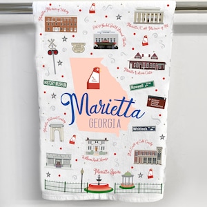May include: A white kitchen towel with a colorful illustration of landmarks in Marietta, Georgia. The illustration includes the Marietta History Museum, Marietta National Cemetery, William Root House, Marietta Square, and the Earl and Rachel Smith Strand Theatre. The text "Marietta, Georgia" is written in blue cursive.