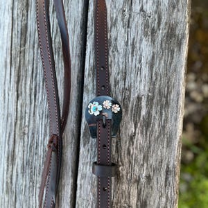 May include: A brown leather strap with a decorative buckle featuring a black background and three colorful flowers. The strap is stitched with a decorative pattern.