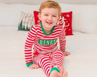 May include: A child wearing red and white striped pajamas with the name "HENRY" on the chest, smiling. The pajamas have green trim. The background includes pillows, Christmas decorations, and a white bed.