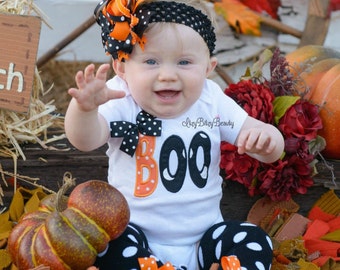 May include: A baby wearing a white onesie with the word "BOO" and the number "00" in orange and black, surrounded by fall decorations. The baby is wearing a black headband with an orange and black bow. Pumpkins and fall leaves are in the background.