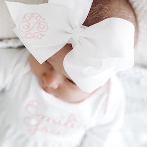May include: A large white hair bow made of grosgrain ribbon featuring a pink embroidered monogram on one side. The bow is attached to a matching white headband. The fabric has a smooth texture and the embroidery consists of cursive letters.