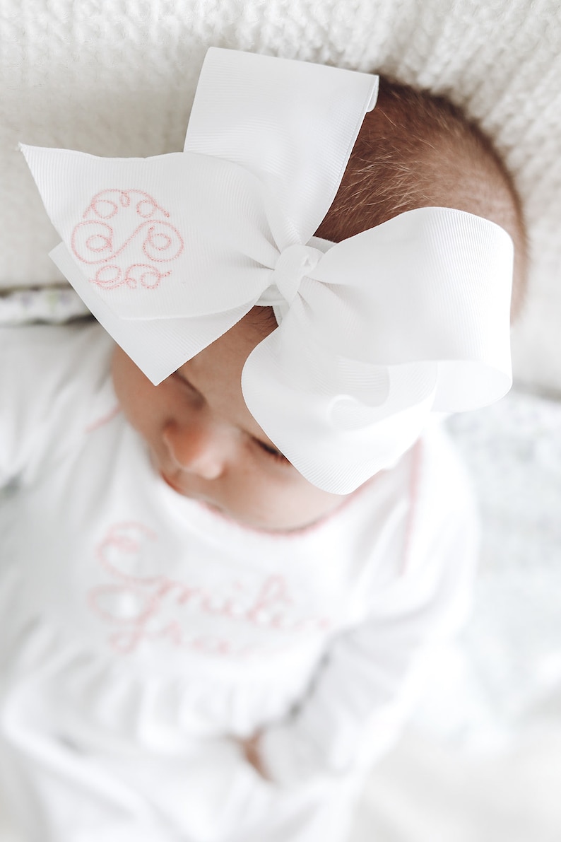 May include: A large white hair bow made of grosgrain ribbon featuring a pink embroidered monogram on one side. The bow is attached to a matching white headband. The fabric has a smooth texture and the embroidery consists of cursive letters.