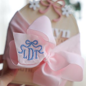 May include: A light pink ribbon bow with embroidered blue initials "LDT" is wrapped around a light-coloured wooden plaque. The bow is accented with a smaller pink bow and white flower embellishments. The background is blurred.