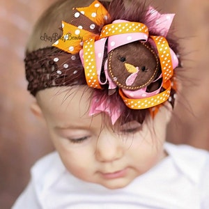 May include: A brown crochet headband adorned with a turkey-themed bow. The bow features orange and pink ribbons with white polka dots, a felt turkey face, and brown feathers. The baby is wearing a white onesie with the name "Brooklynn's" in pink.