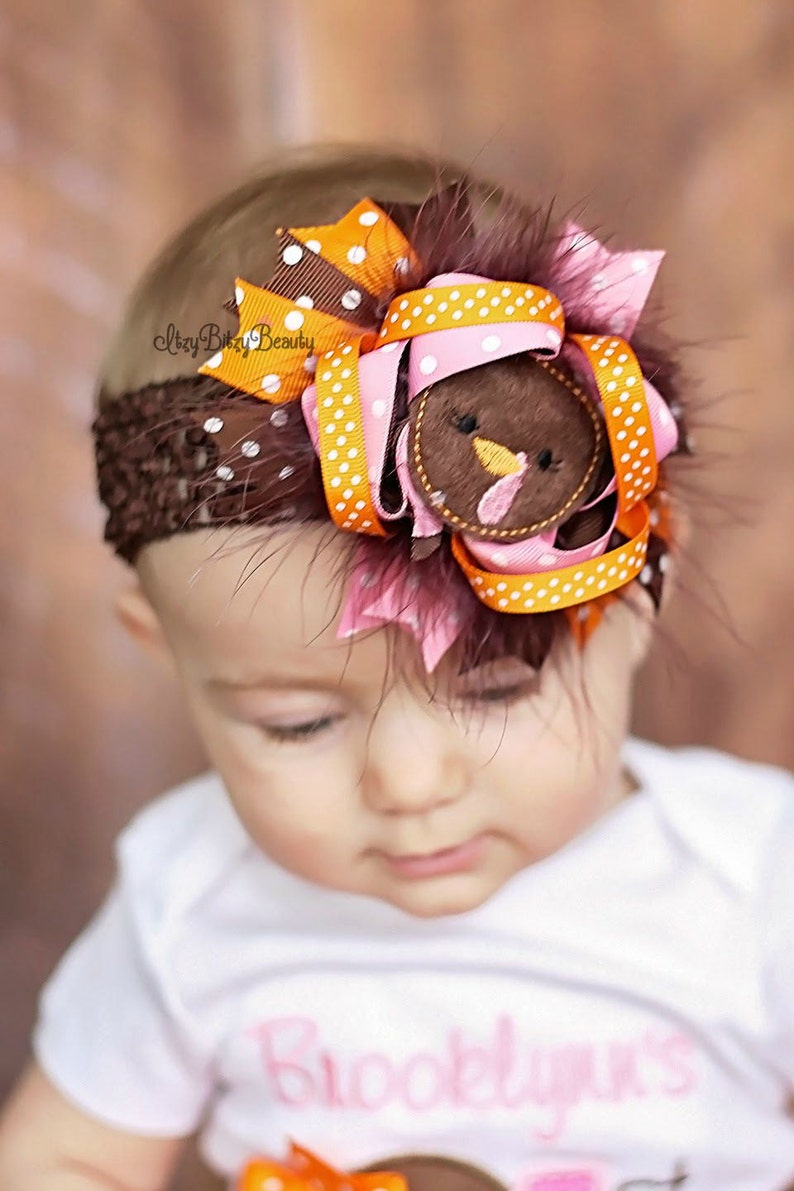 May include: A brown crochet headband adorned with a turkey-themed bow. The bow features orange and pink ribbons with white polka dots, a felt turkey face, and brown feathers. The baby is wearing a white onesie with the name "Brooklynn's" in pink.