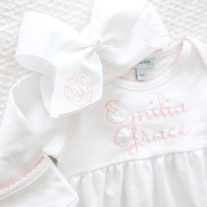 May include: A white long-sleeved bodysuit with a gathered waist and pink scalloped trim on the cuffs and neckline. The name Emilia Grace is embroidered in pink on the chest. A large white hair bow with a pink embroidered monogram is included.