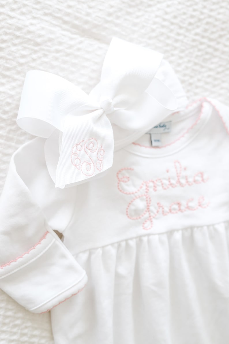 May include: A white long-sleeved bodysuit with a gathered waist and pink scalloped trim on the cuffs and neckline. The name Emilia Grace is embroidered in pink on the chest. A large white hair bow with a pink embroidered monogram is included.