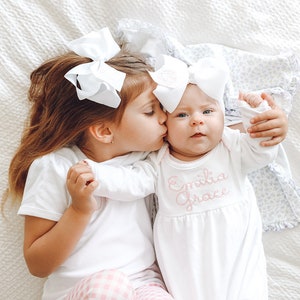May include: A white short-sleeved shirt and a white long-sleeved gown with the embroidered name Emilia Grace in pink script. Both garments are paired with large white hair bows. The gown features a gathered waist and a simple round neckline.