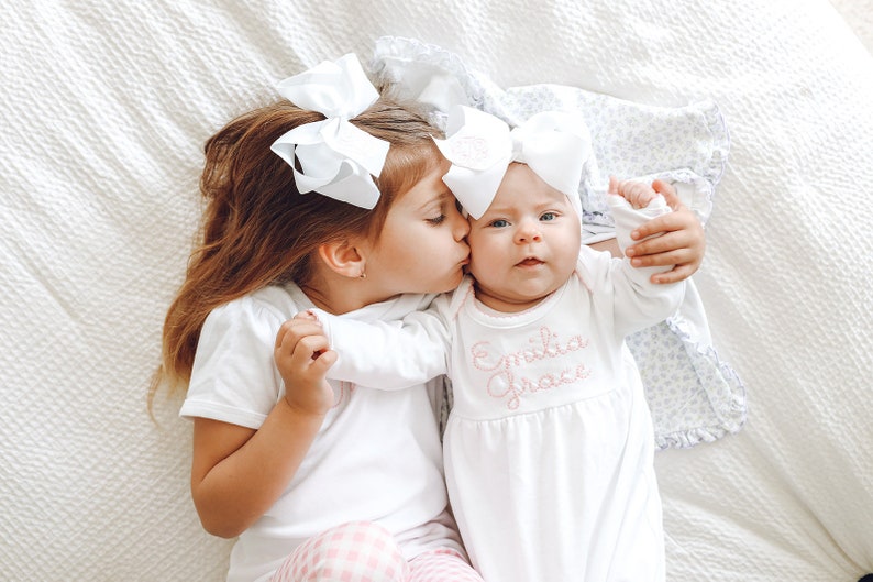 May include: A white short-sleeved shirt and a white long-sleeved gown with the embroidered name Emilia Grace in pink script. Both garments are paired with large white hair bows. The gown features a gathered waist and a simple round neckline.