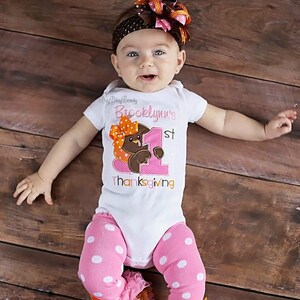 May include: A baby wearing a white onesie with the text "Brooklynn's 1st Thanksgiving" and a turkey graphic. The baby is also wearing pink polka dot leggings with ruffles and orange bows, and a brown headband with a large bow.