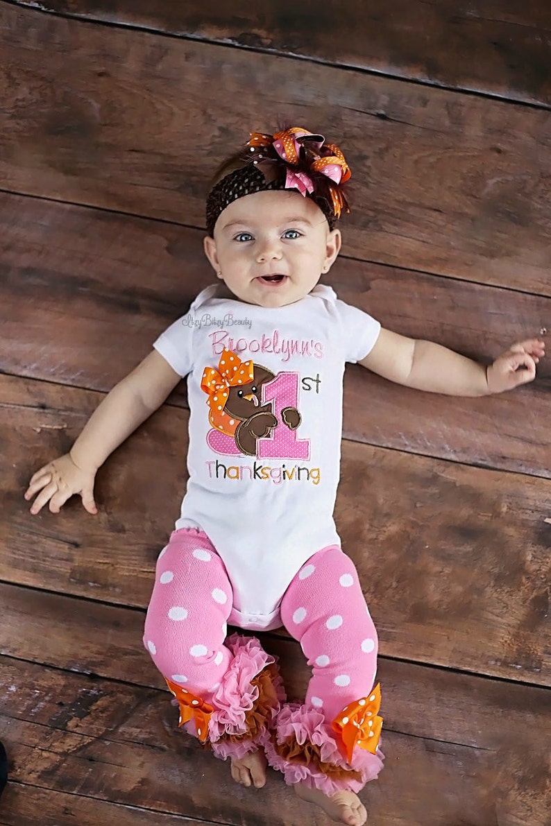 May include: A baby wearing a white onesie with the text "Brooklynn's 1st Thanksgiving" and a turkey graphic. The baby is also wearing pink polka dot leggings with ruffles and orange bows, and a brown headband with a large bow.
