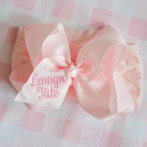 May include: A pale pink baby headband featuring a large bow. The bow is embroidered with the name "Lennyyn Tate" in pink thread. The headband is set against a pink and white gingham background.