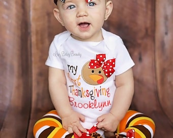 May include: A baby wearing a Thanksgiving outfit, including a white onesie with a turkey applique and the text "My First Thanksgiving Brooklyn". The outfit includes striped leggings and a turkey-themed headband.