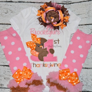 May include: A white onesie with a turkey graphic and the text "Brooklyn's 1st Thanksgiving." The outfit includes pink and white polka dot leg warmers, orange bows, and a matching turkey hair bow. A festive outfit for a baby's first Thanksgiving.