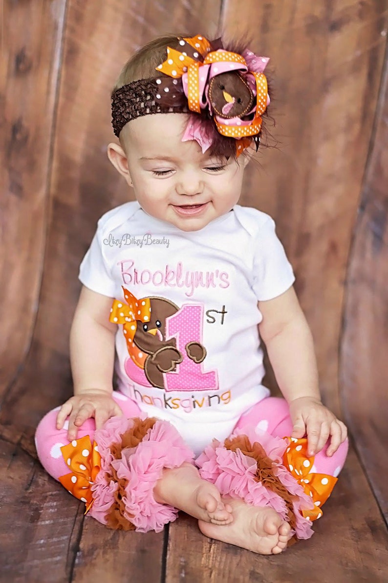 May include: A baby wearing a white onesie with a turkey graphic and the text "Brooklynn's 1st Thanksgiving." The outfit includes pink polka dot pants with ruffles and a matching headband with a turkey and bow.