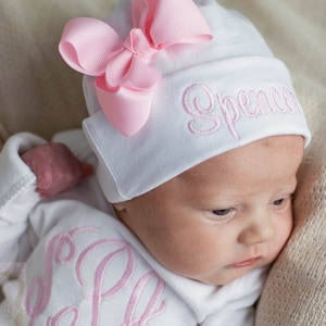 May include: A white baby hat with a pink bow and the name "Spencer" embroidered on the front. The hat is worn by a baby with a white onesie that has the name "Sophia" embroidered on the front.