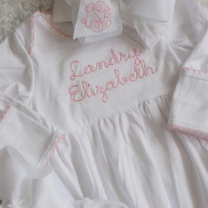 May include: A white long-sleeved gown with pink embroidered stitching along the neckline and cuffs. The name Landry Elizabeth is embroidered in pink on the chest. A matching white hair bow with a pink monogram is positioned at the top.