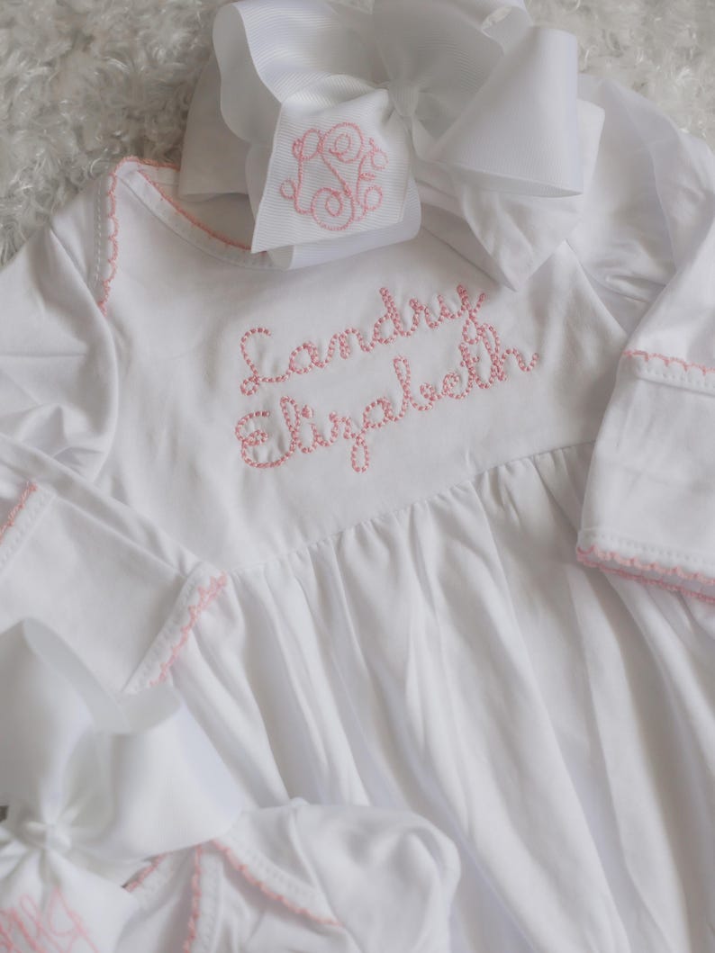 May include: A white long-sleeved gown with pink embroidered stitching along the neckline and cuffs. The name Landry Elizabeth is embroidered in pink on the chest. A matching white hair bow with a pink monogram is positioned at the top.