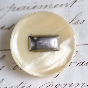 Puede incluir: Un botón blanco con un rectángulo de metal plateado en el centro. El botón tiene un acabado nacarado.