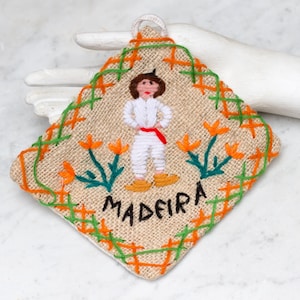 Op de afbeelding: Een vierkant van beige jute met groene en oranje borduurwerk. In het midden is een figuur in een wit shirt en een rode sjerp, met het woord "MADEIRA" eronder geborduurd.