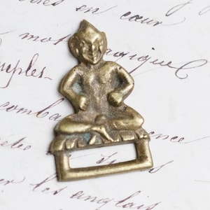 Op de afbeelding: Een klein, messing beeldje van een zittend figuur met een puntige hoed en een ondeugende uitdrukking. Het beeldje is bevestigd aan een rechthoekige lus, wat suggereert dat het een gesp of een decoratieve sluiting kan zijn.