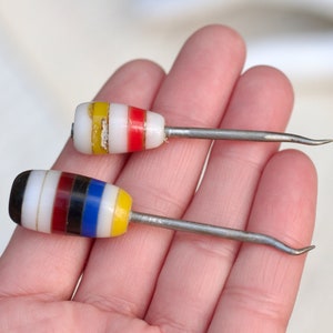 Puede incluir: Dos pequeñas herramientas con puntas de vidrio de colores y mangos de metal. Las puntas están rayadas de rojo, blanco, azul, negro y amarillo.