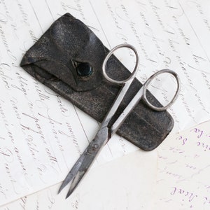 Puede incluir: Un par de tijeras de plata antiguas con una funda de cuero. Las tijeras tienen un diseño sencillo con mangos redondeados y una punta puntiaguda. La funda de cuero está desgastada y tiene un color marrón oscuro.