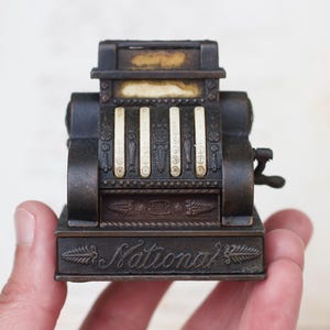 May include: A vintage metal National cash register pencil sharpener with a black and brown finish. The sharpener has a lever on the side and a drawer for shavings.
