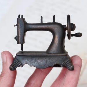 May include: A small, antique-looking metal sewing machine pencil sharpener with a black finish and intricate details. The sharpener has a hand crank and a decorative base.