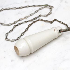 Könnte beinhalten: Antiker Toiletten-Zuggriff aus weißem Porzellan mit Metallkette. Der Griff ist zylindrisch mit abgerundetem Ende und weist Altersspuren auf. Die Kette besteht aus kleinen, ineinandergreifenden Metallgliedern.