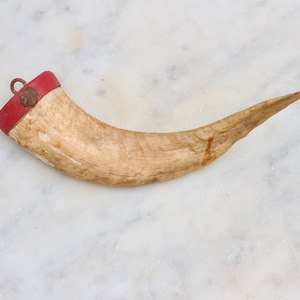 Pode incluir: Um chifre de madeira com uma borda vermelha e um laço de metal no topo. O chifre é de cor castanho claro e tem um acabamento natural.