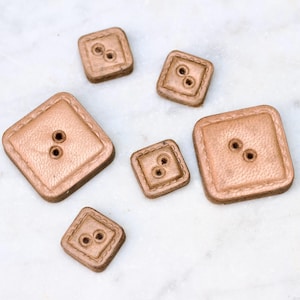 May include: Five square-shaped leather buttons in various sizes. The buttons are a light brown color and have two holes each.