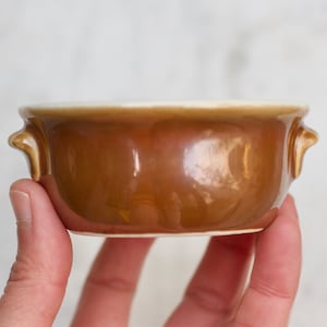 May include: A small brown ceramic bowl with two handles. The bowl is glazed and has a slight sheen.