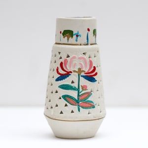 May include: A cream-coloured ceramic vase with a floral design. The vase features a pink and red flower with green and blue leaves. The vase has a geometric pattern of small triangles and is accented with gold trim.