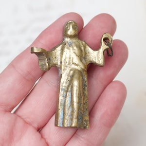 Puede incluir: Una pequeña figurita de metal dorado de una persona con los brazos extendidos. La figurita tiene un lazo en la parte superior para colgar.