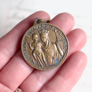 Puede incluir: Una medalla de bronce con una representaci&oacute;n de la Virgen Mar&iacute;a y el ni&ntilde;o Jes&uacute;s. La medalla est&aacute; inscrita con las palabras "Ave Maria, B.V.M. Moeder".