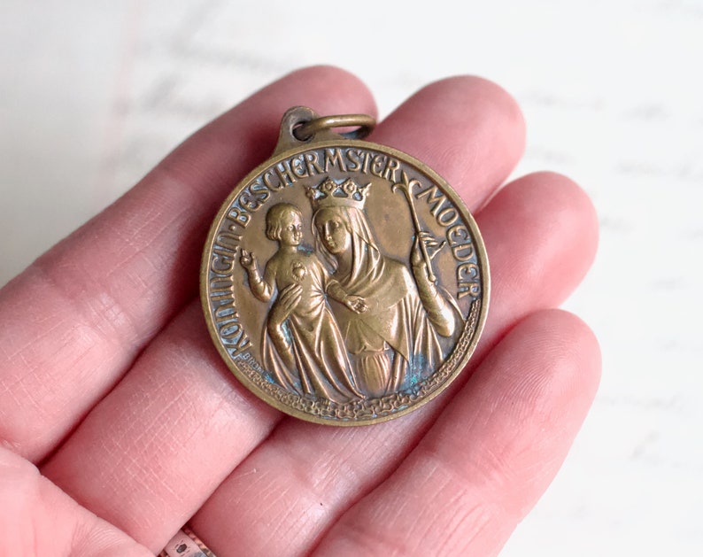 Puede incluir: Una medalla de bronce con una representaci&oacute;n de la Virgen Mar&iacute;a y el ni&ntilde;o Jes&uacute;s. La medalla est&aacute; inscrita con las palabras "Ave Maria, B.V.M. Moeder".