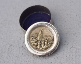 Puede incluir: Una caja de metal plateado con interior de terciopelo azul. La tapa presenta una imagen en relieve de una escena religiosa con una mujer y varias figuras.
