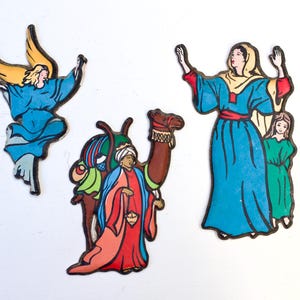 Può includere: Tre addobbi natalizi vintage che raffigurano i tre re magi. Il primo addobbo è un angelo blu con capelli e ali gialli. Il secondo addobbo è un uomo che cavalca un cammello con una tunica rossa e gialla. Il terzo addobbo è una donna con una tunica blu con un foulard giallo in testa e una bambina con un vestito verde.