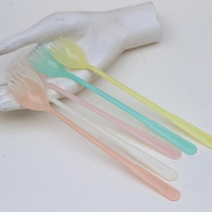 Puede incluir: Cinco tenedores de postre de plástico de color pastel con una cuchara en el extremo. Los tenedores son amarillos, azul claro, rosas, blancos y rosa claro.