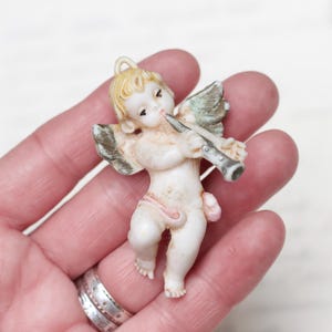 Puede incluir: Una figurita de ángel de porcelana blanca tocando la flauta. El ángel tiene el pelo castaño claro y una faja rosa. La figurita sostiene la flauta en su mano derecha y tiene su mano izquierda en la cadera.