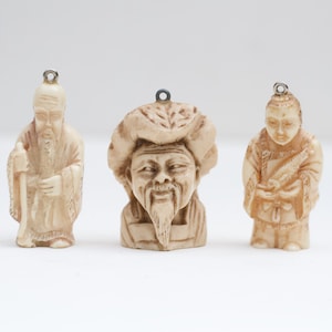Peut inclure: Trois petites figurines en ivoire de couleur crème représentant des hommes asiatiques avec des traits du visage détaillés et des vêtements traditionnels. Les figurines sont des pendentifs avec une boucle en haut pour les suspendre.