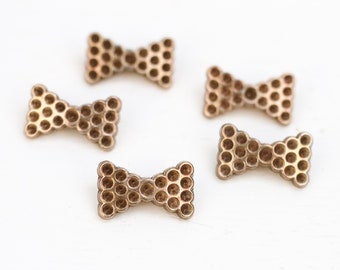 May include: Close-up of several small, bow-tie shaped embellishments. The bows are a metallic gold color and feature a pattern of small, evenly spaced holes. They are scattered on a white surface.